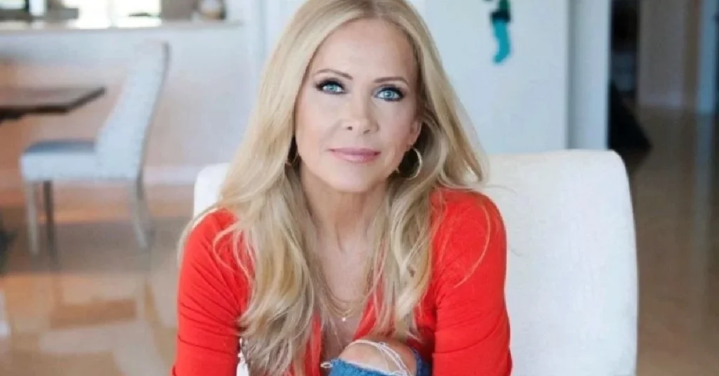 Shemane Deziel sitting on a white chair in a modern living room, wearing a red top and jeans.